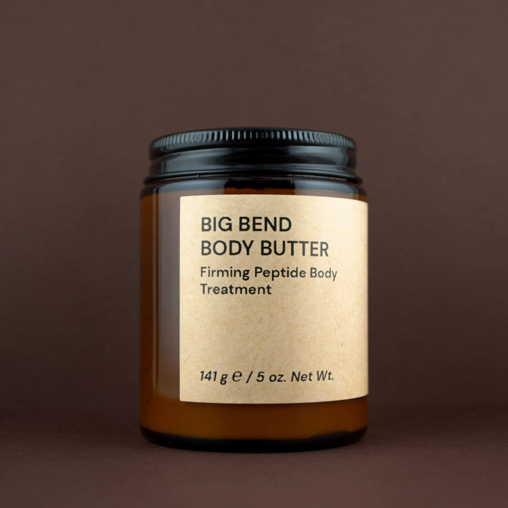 A jar of Big Bend Body Butter Firming Peptide Body Treatment, featuring an amber glass jar with a black lid and a beige kraft paper label, set against a solid dark brown background.